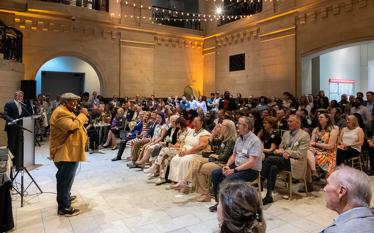 A smiling Black man speaks to a large group of people seated in the Great Hall